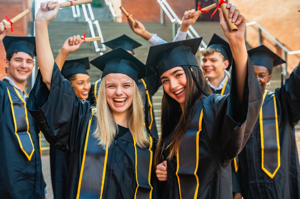 Diverse students wearing graduation caps and gowns are holding up their diplomas, cheering happily