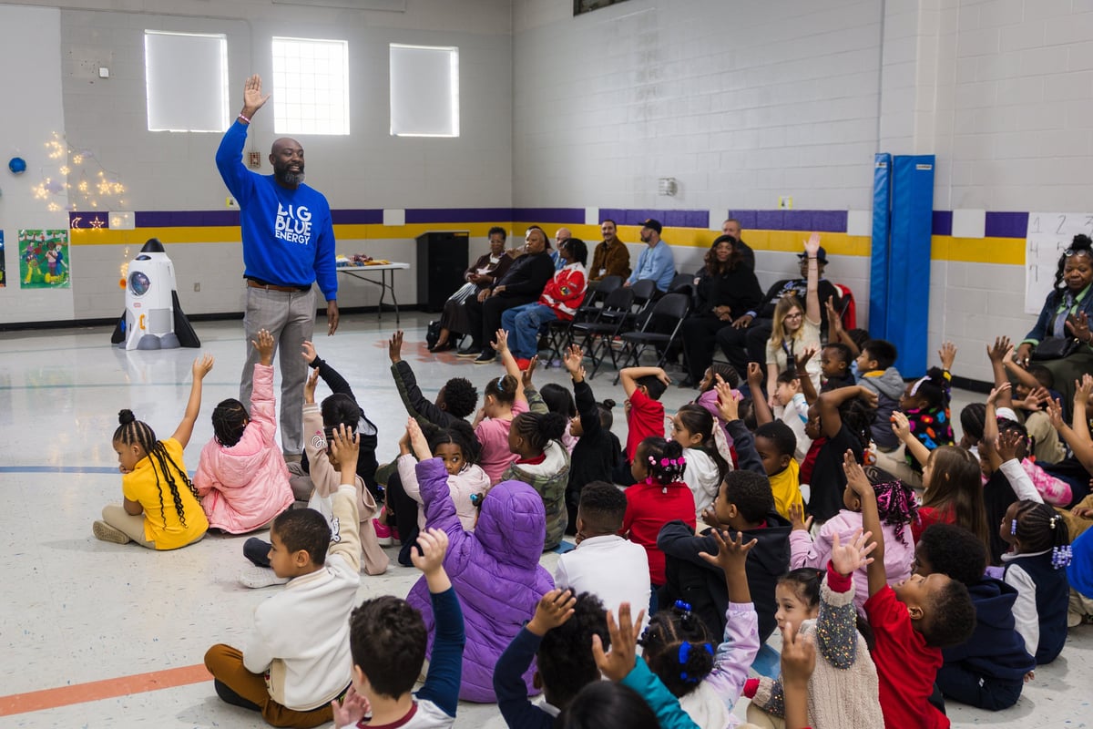 An instructor in a blue shirt leads an interactive activity with a large group of young children sitting on a gymnasium floor during a school event